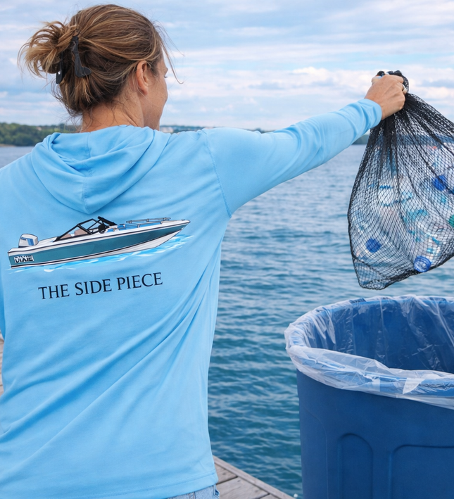 Person in a blue hoodie with 'The Side Piece' logo, holding a fishing net over a trash bin by a body of water.